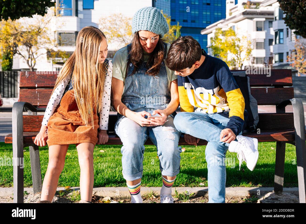 Mother with kids using smart phone sitting on bench in public park ...