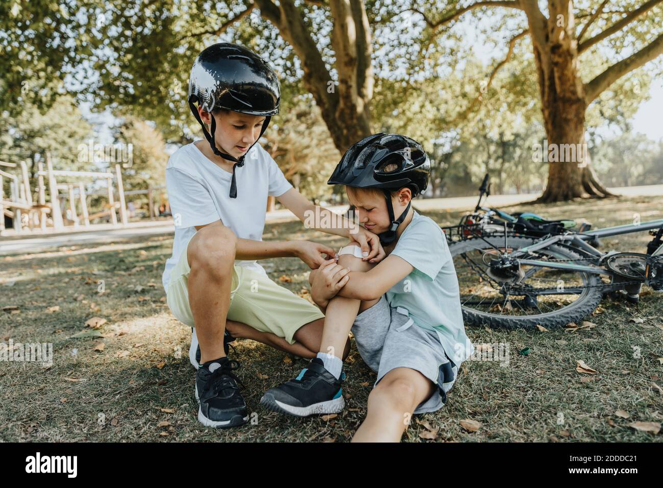 Boy putting bandage on younger brother knee sitting in public park Stock Photo