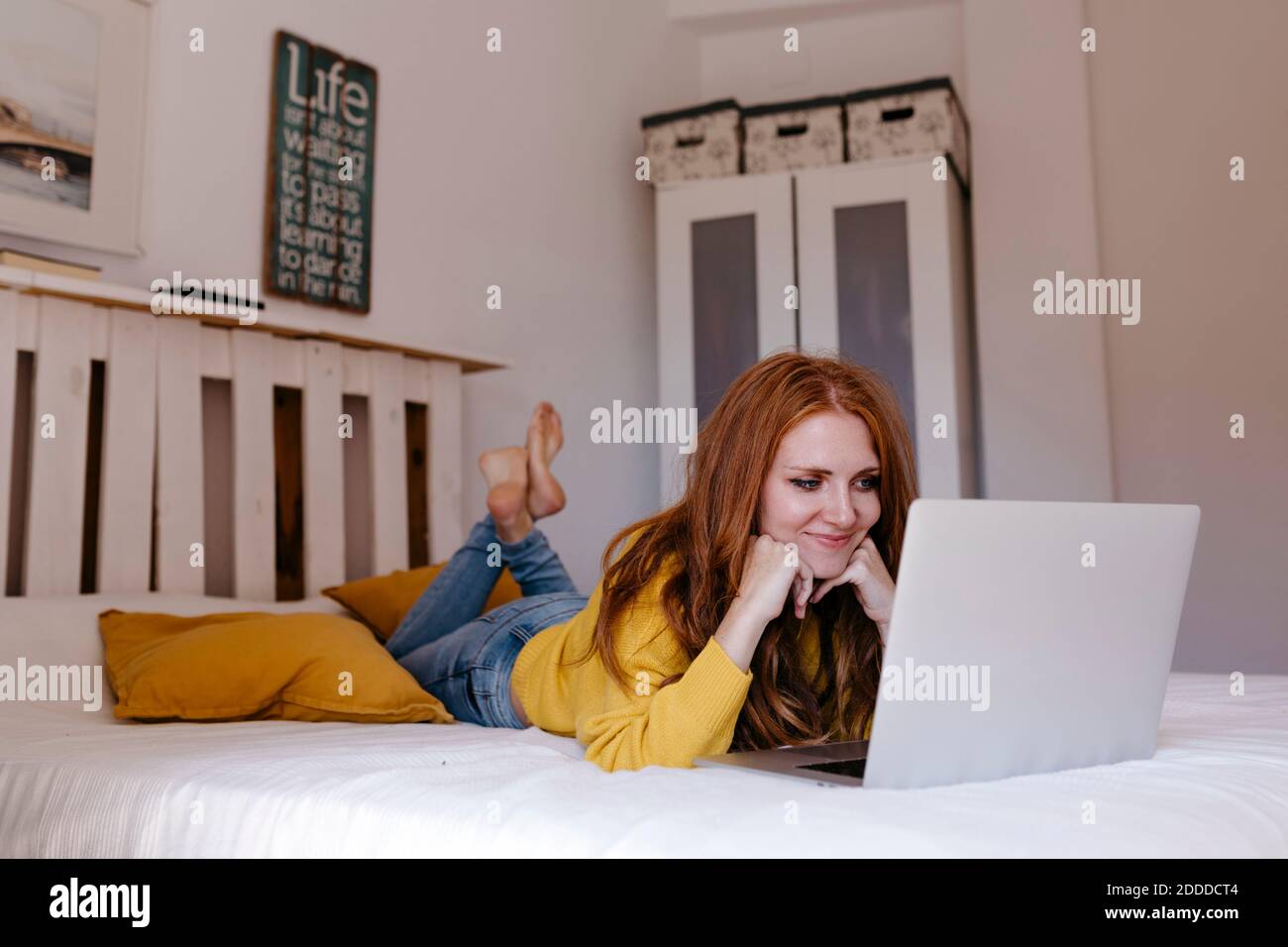 Smiling woman lying on front with hand on chin using laptop at home Stock Photo