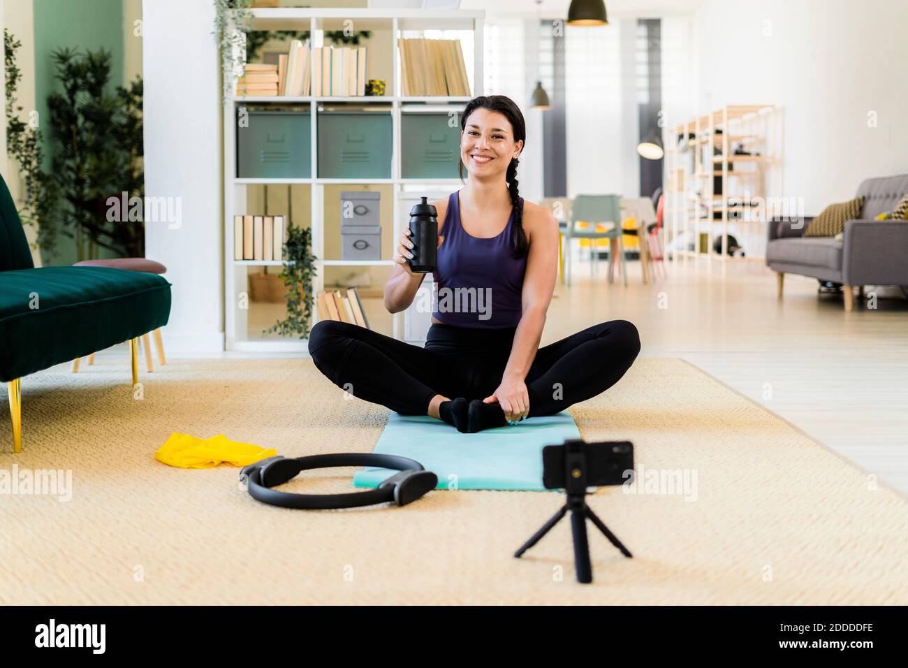 Young woman smiling with bottle video recording on camera while sitting in cobbler pose at home Stock Photo