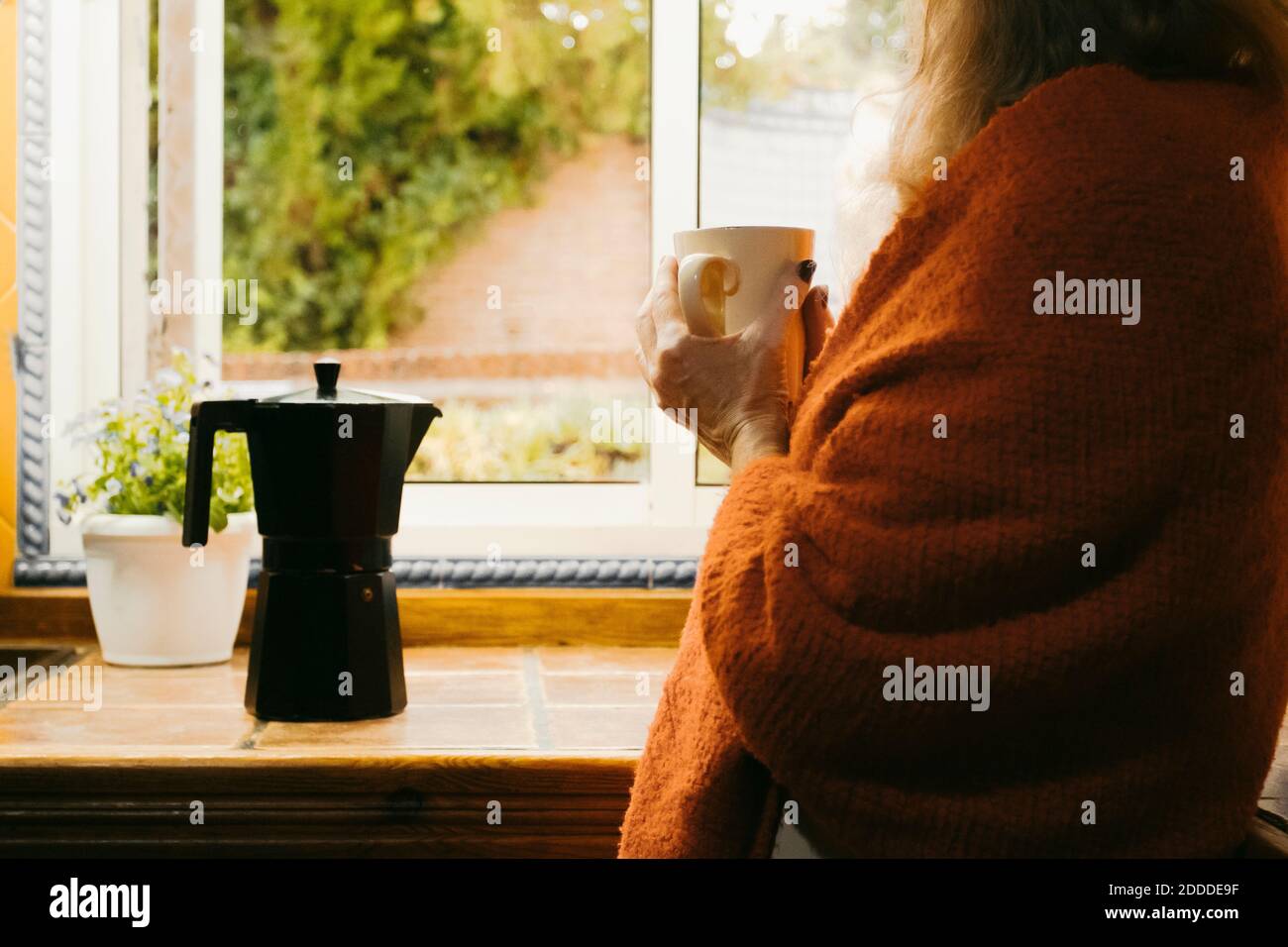 Woman wrapped in a blanket drinking coffee while standing by window in kitchen at home Stock Photo