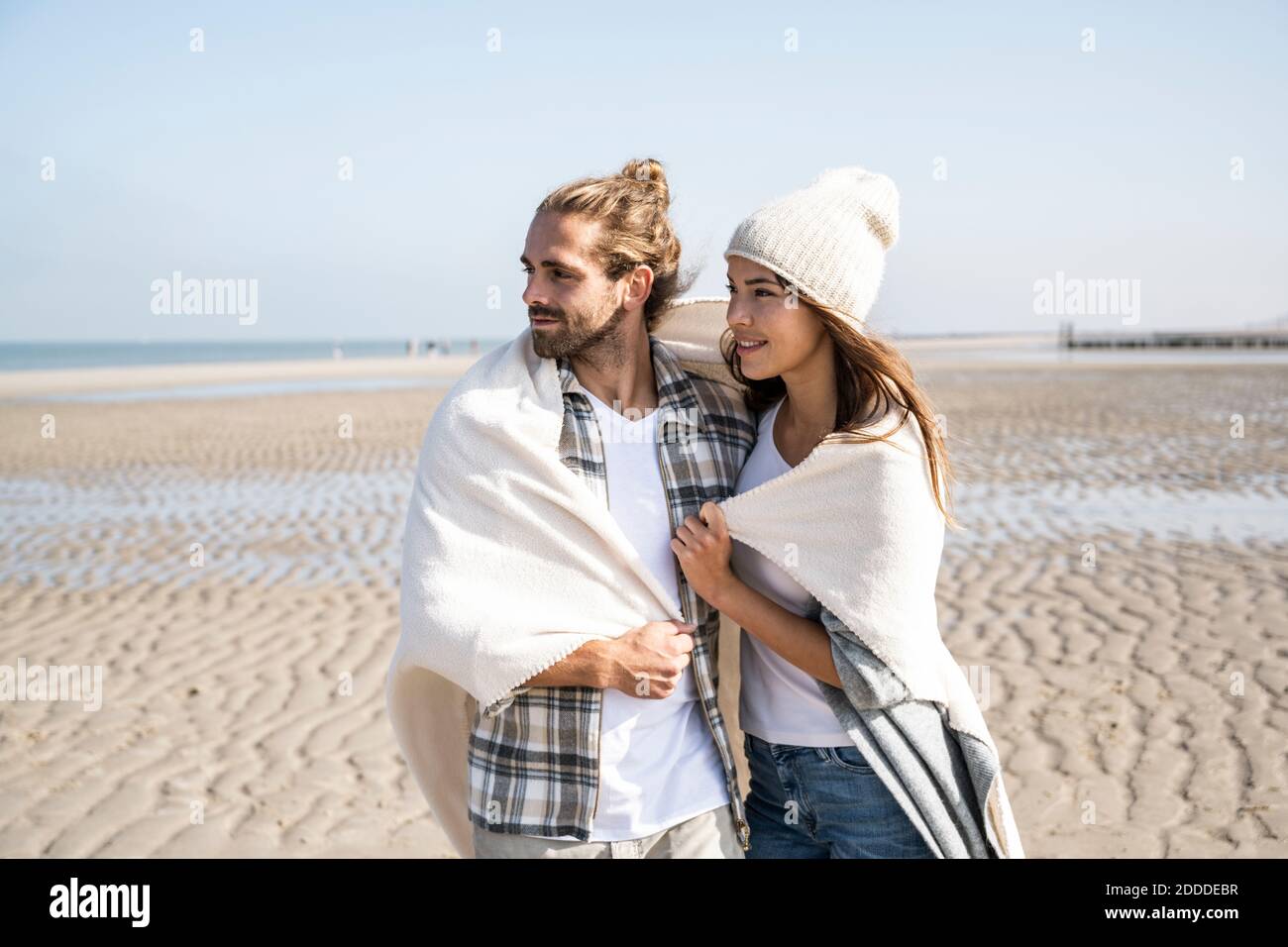 Thoughtful young couple covered in blanket looking away while standing at beach on sunny day Stock Photo