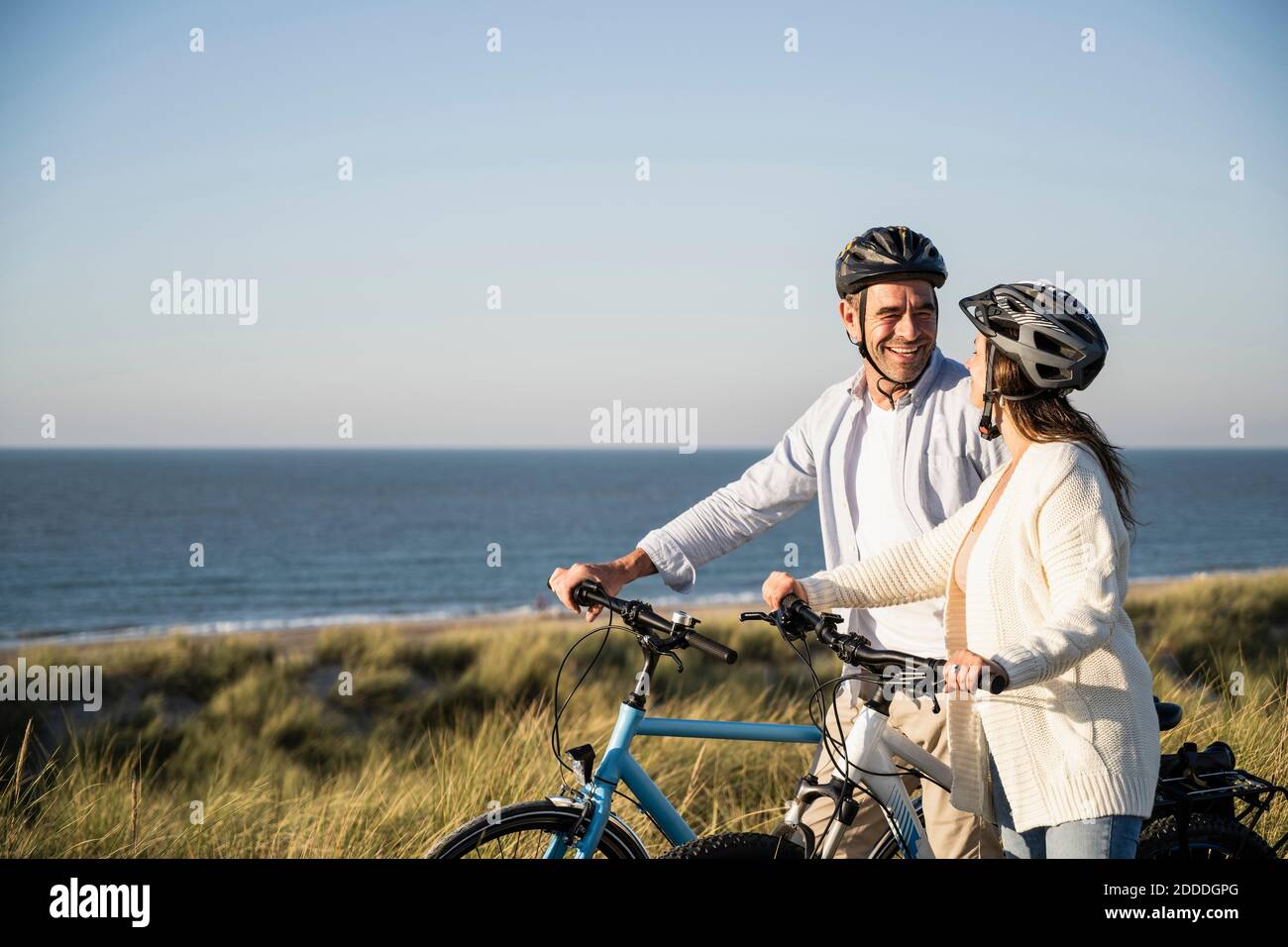 Happy couple in cycling helmets standing with bicycle at beach against clear sky Stock Photo