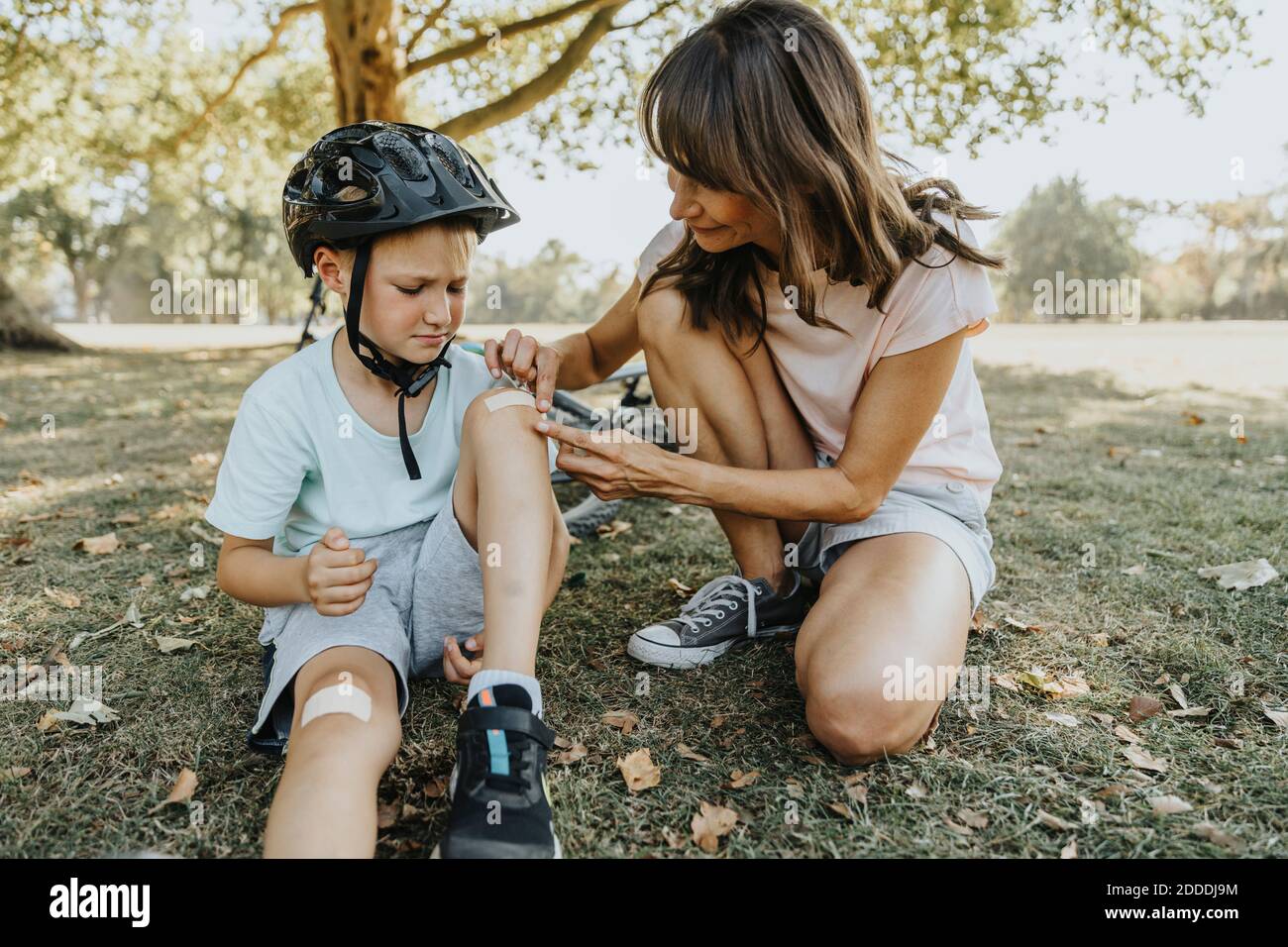 Mother putting bandage on son's knee while sitting in public park during sunny day Stock Photo