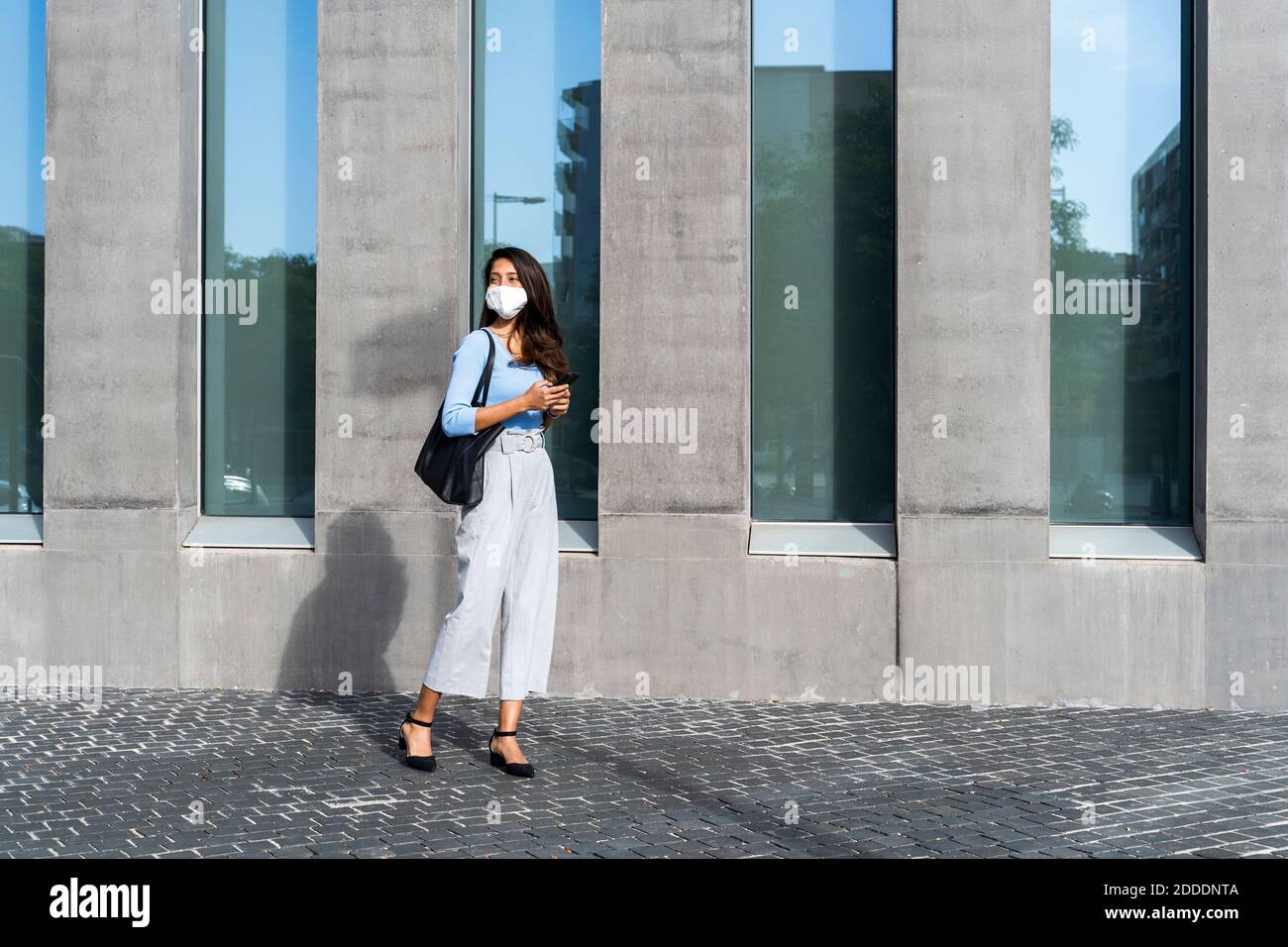 Businesswoman in face mask standing on footpath by building during coronavirus crisis Stock Photo