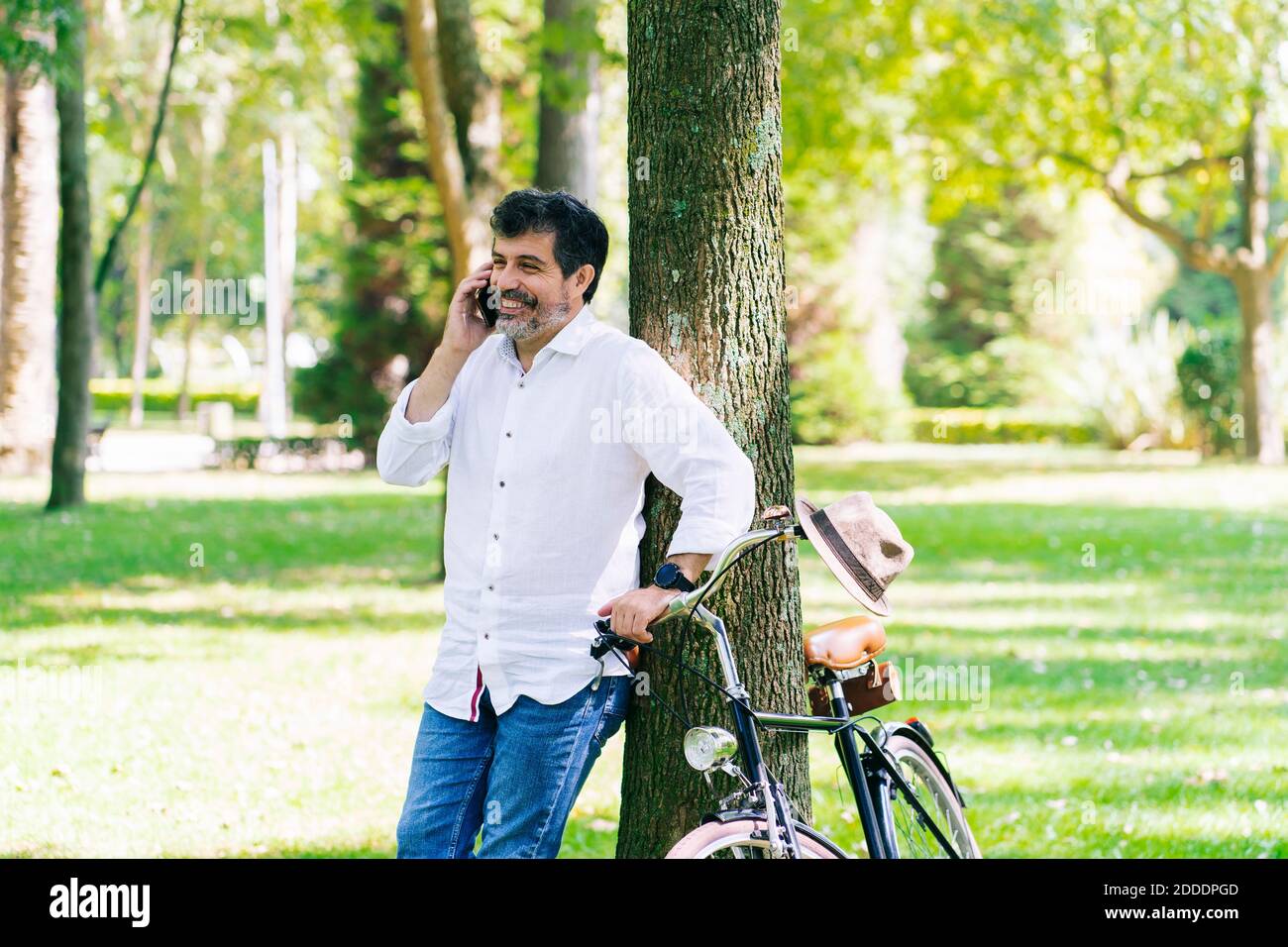 Happy mature man talking on smart phone while leaning on tree at public park Stock Photo