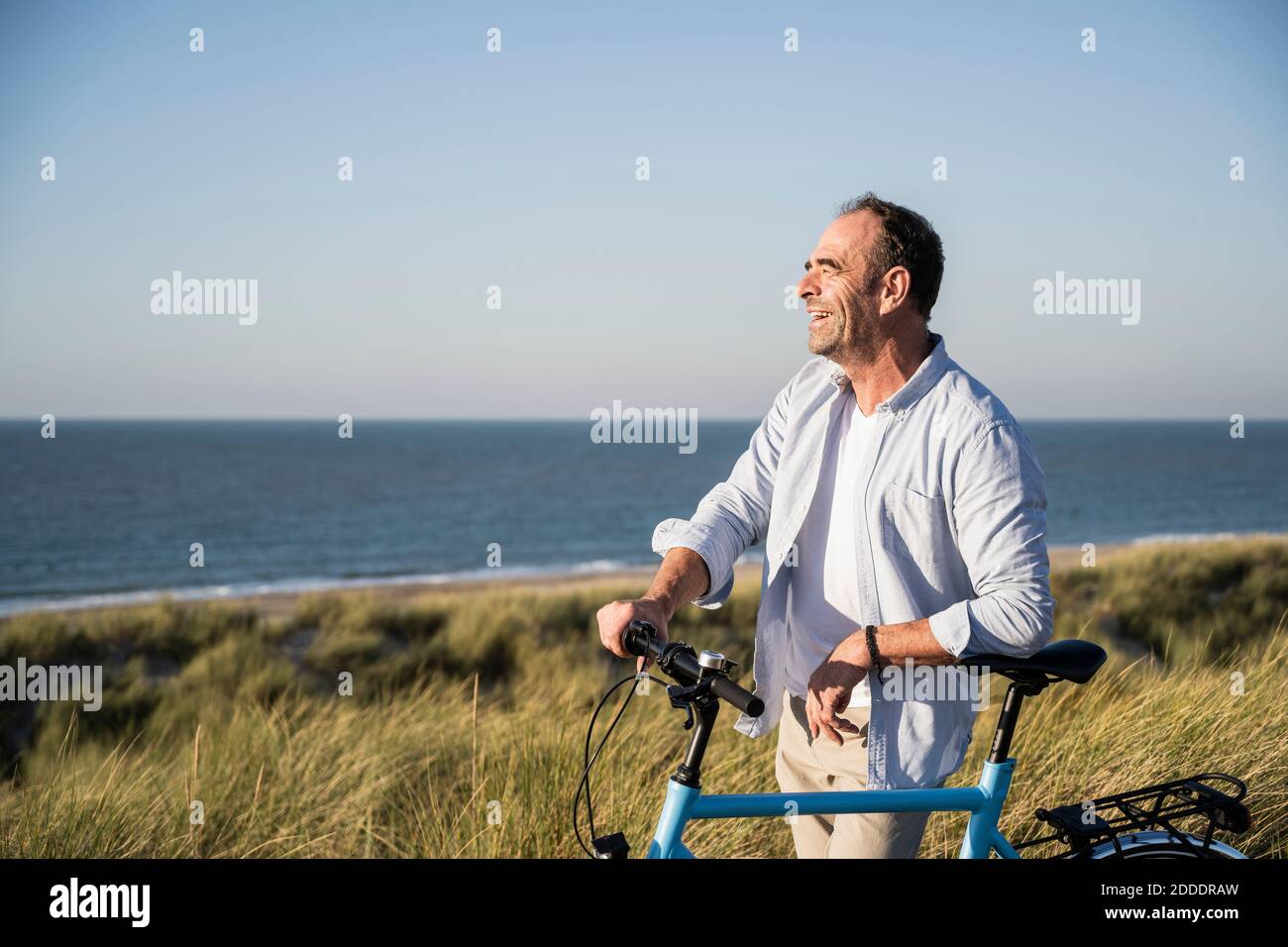 Cheerful mature man looking away while standing with bicycle at beach against clear sky Stock Photo
