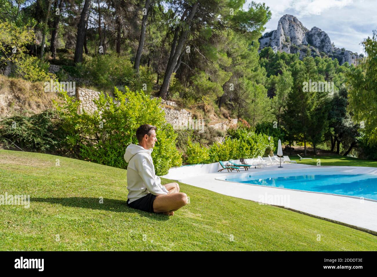 Mature man meditating while practicing yoga on grass near swimming pool at health retreat during sunny day Stock Photo