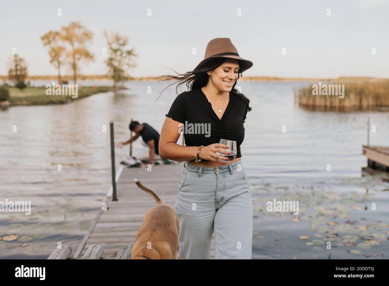 Smiling woman holding wineglass while walking with dog and man in background on pier Stock Photo