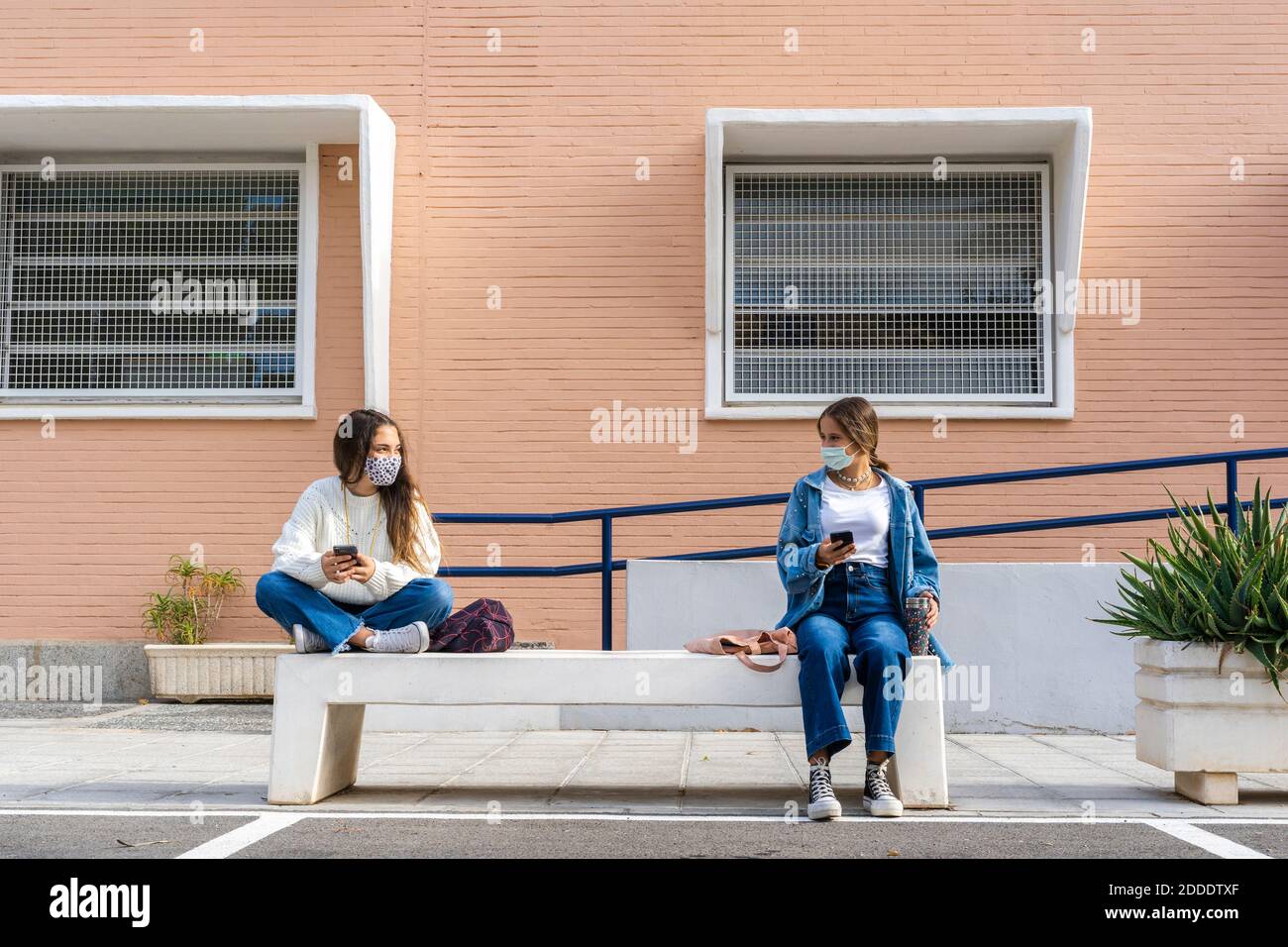 Female friends social distancing wearing protective face mask sitting on concrete bench Stock Photo
