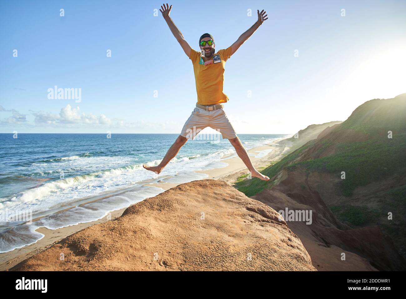 Cheerful man jumping on rock formation at beach against blue sky Stock Photo