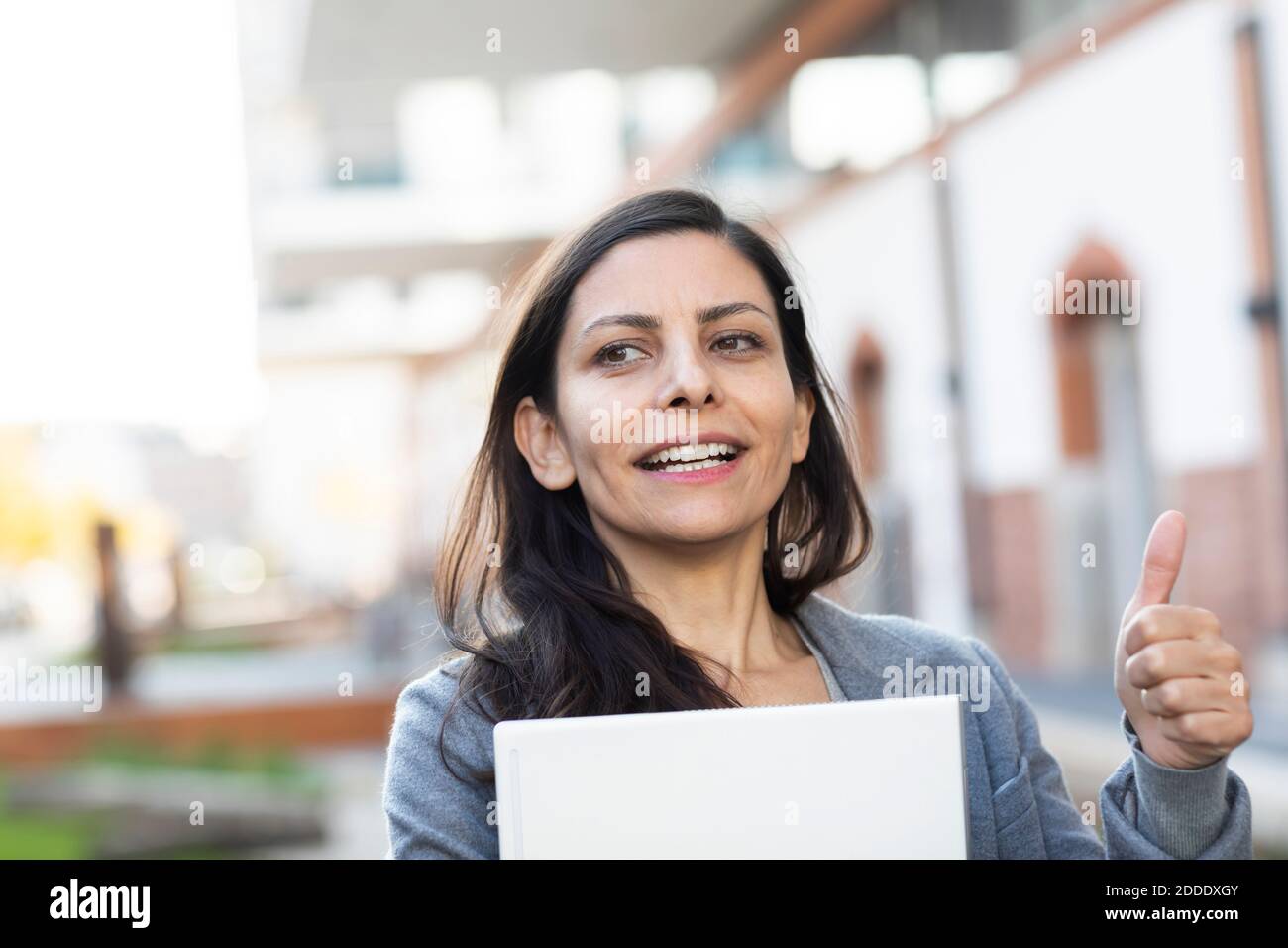 Businesswoman with laptop showing thumps up while standing against building Stock Photo