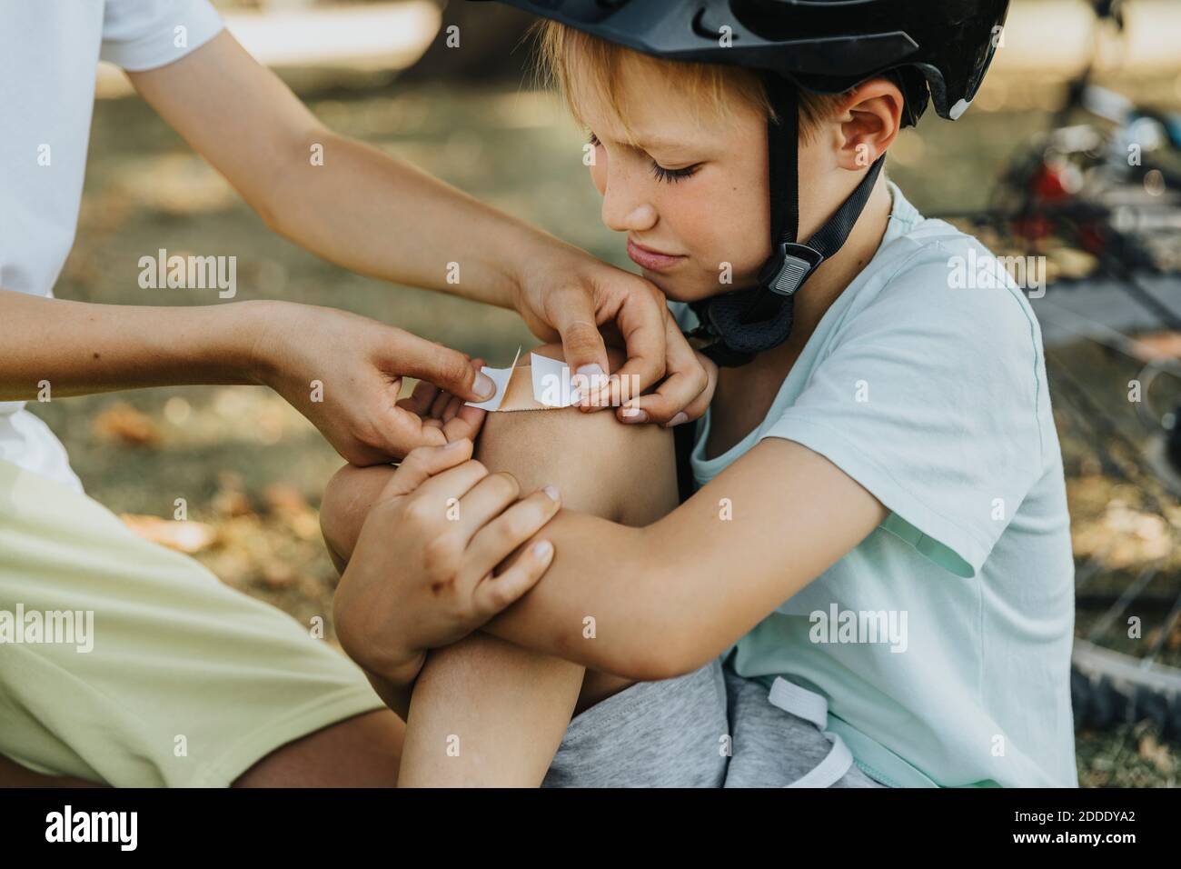 Boy putting bandage on younger brother knee sitting in public park Stock Photo