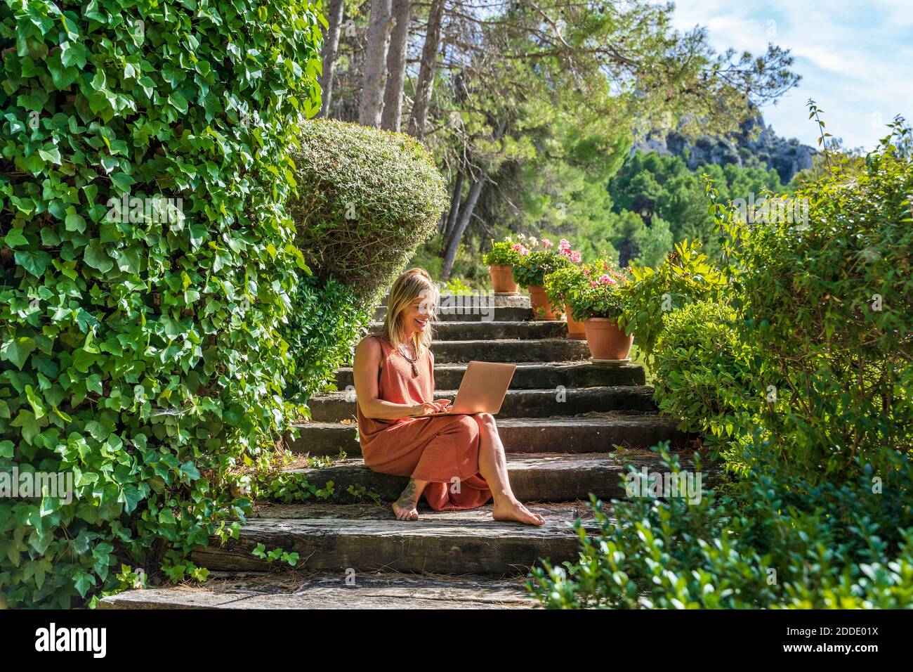 Mature woman using laptop while sitting on steps amidst green plants at health retreat during sunny day Stock Photo