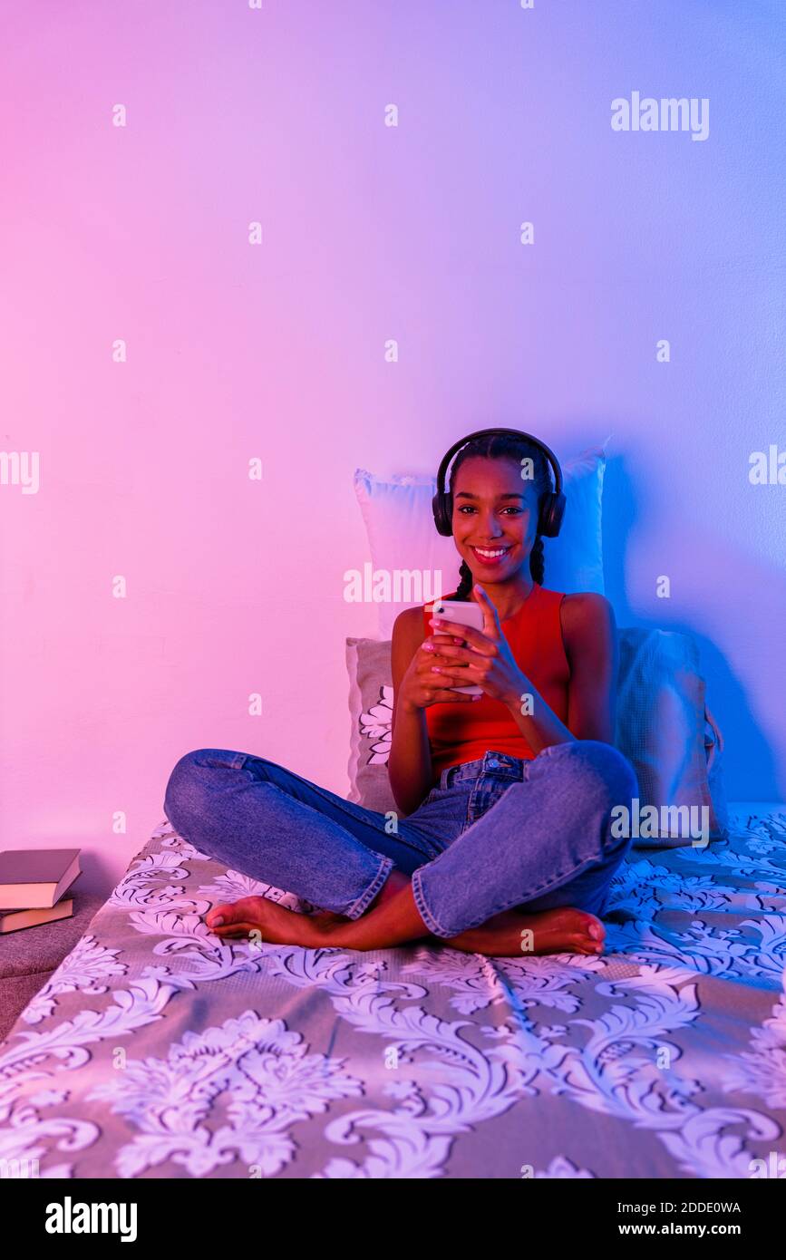 Smiling teenage girl using smart phone sitting on bed at home in ...