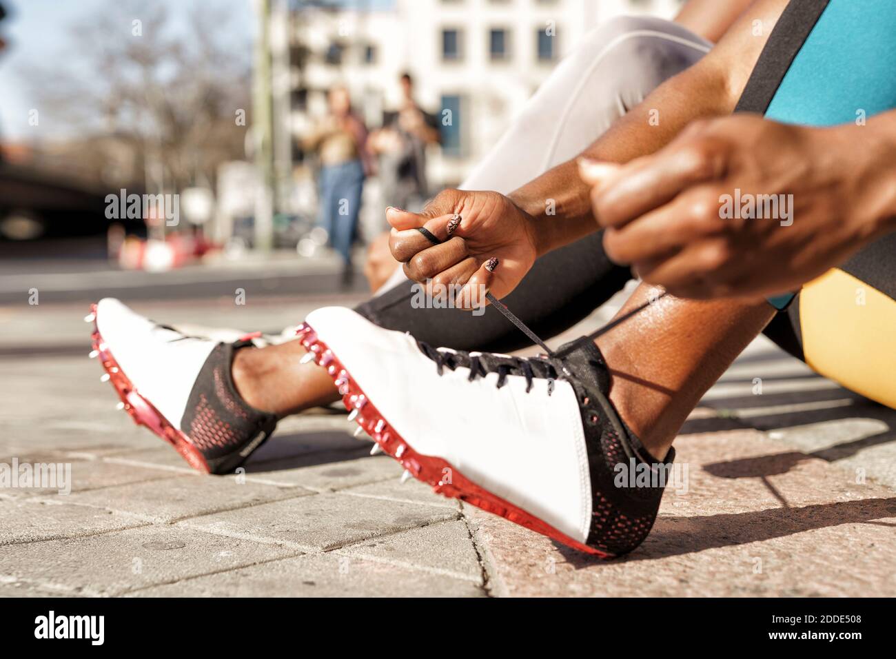 Sportswoman typing shoelace while sitting by man on sidewalk at city Stock Photo