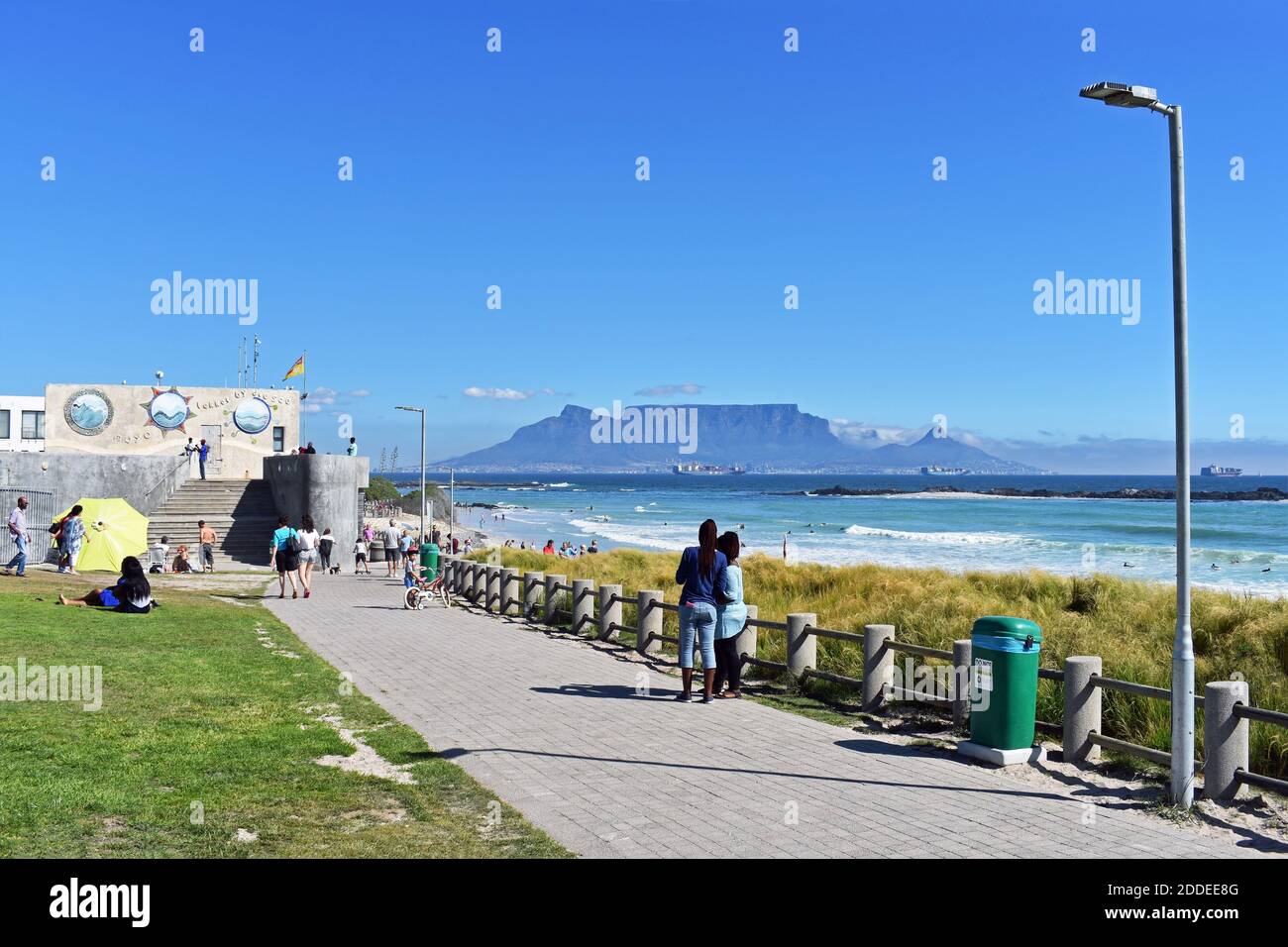 Table Mountain, Devils Peak & Lions Head seen from Bloubergstrand Beach across Table Bay. People walk along the beachside path and sit on the grass. Stock Photo