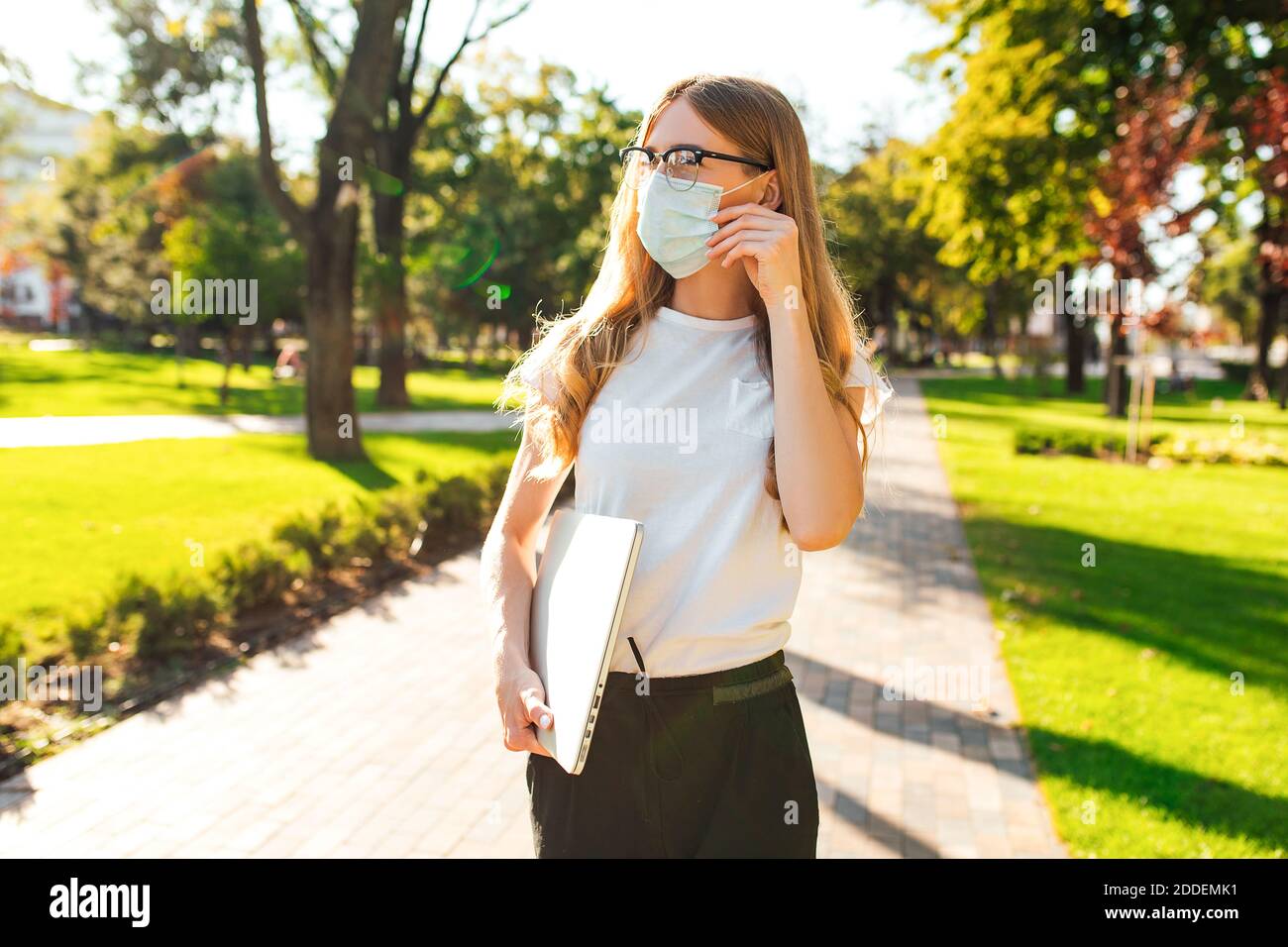 Young attractive woman in a medical protective mask on her face walks along a city street with a laptop, coronavirus concept Stock Photo