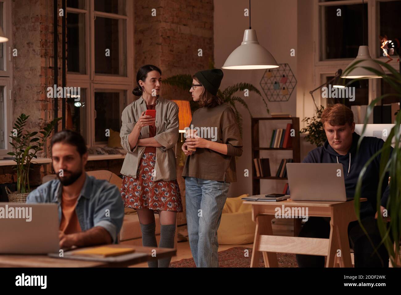 Two young women talking together while standing at office with their colleagues working at their workplaces Stock Photo