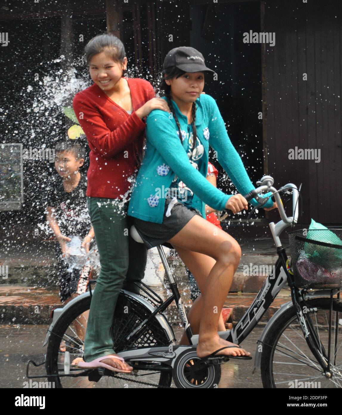 Two young girls on a bike being soaked by water as they pass a young boy enjoying the Songkran Water throwing festival. Stock Photo