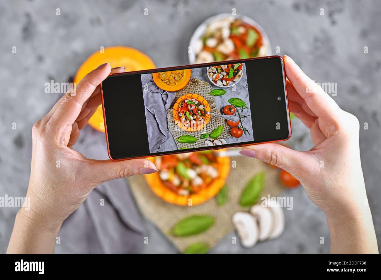 Taking photo of food with hands holding mobile phone with picture of vegan baked Red kuri squash vegetable filled with bell pepper, tomatoes and mushr Stock Photo