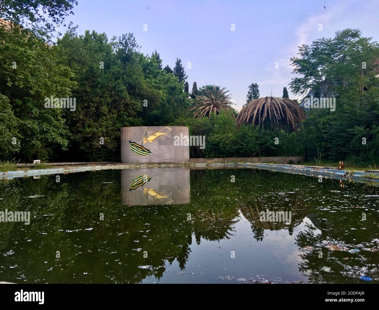 Abandoned dirty public swimming pool surrounded by green tall trees. Trees and wall reflection on the dirty green waters. Fallen autumn leaves. Stock Photo