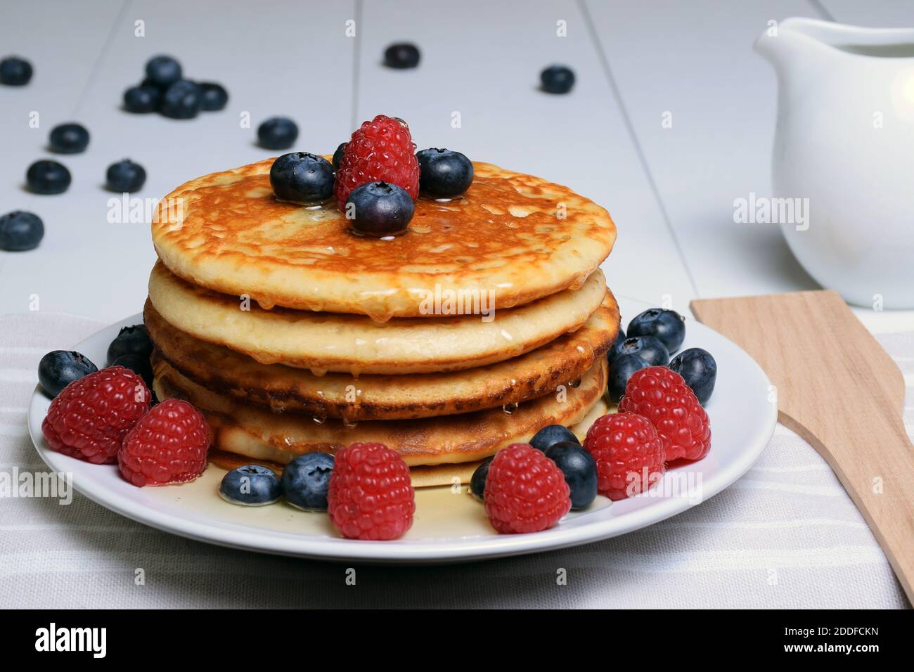 Pancakes with maple syrup, blueberries and raspberries on white wooden table, porcelain jug and wooden spoon. Stock Photo