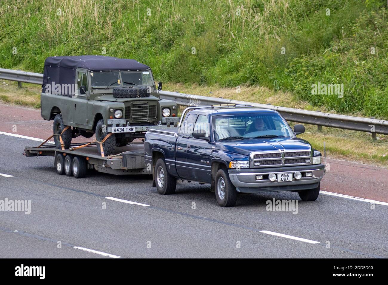 Left hand drive 2000 American Chrysler SUV pickup truck towing 1983 green Land Rover 109' - 6 CYL on trailer; Vehicular traffic, moving vehicles, cars, vehicle driving on UK roads, USA motors, motoring on the M6 motorway highway UK road network. Stock Photo