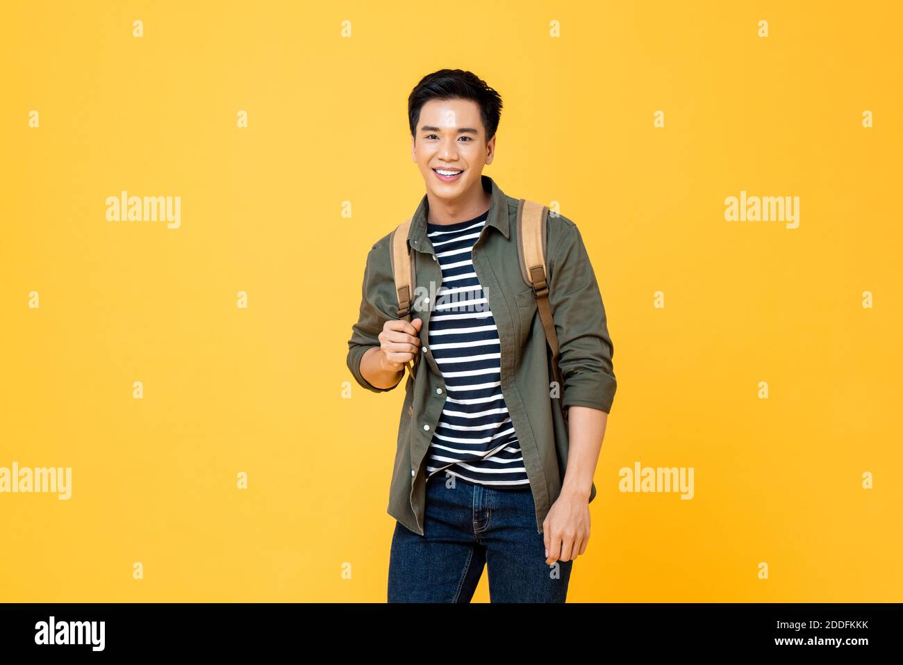 Portrait of smiling young male Asian tourist carrying backpack ready to travel in isolated studio yellow background Stock Photo