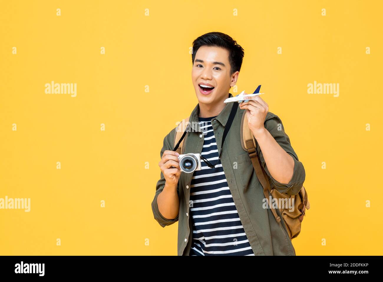 Portrait of young handsome smiling Asian male tourist backpacker holding plane model and camera isolated on studio yellow background Stock Photo