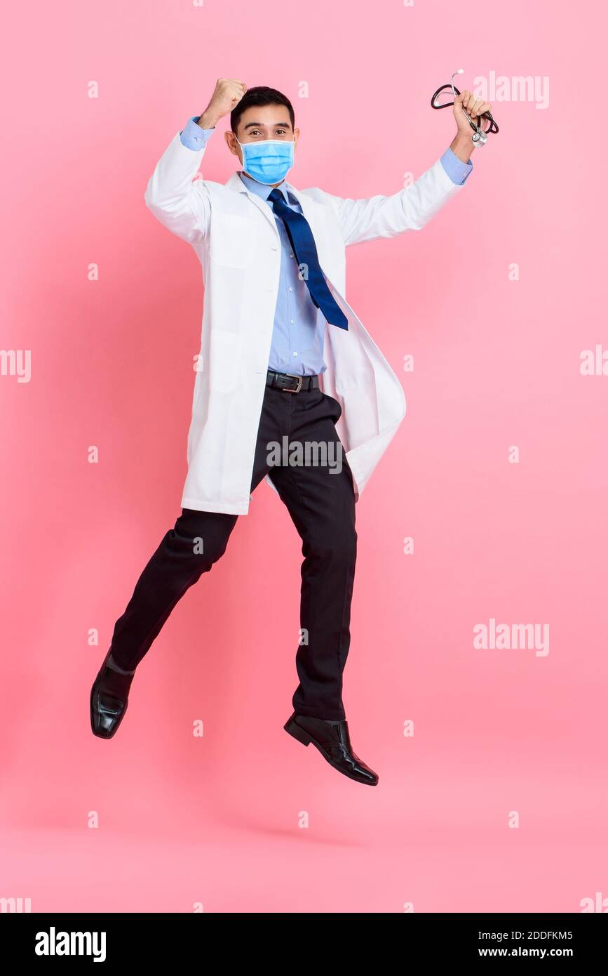 Asian male doctor wearing medical mask raising fists and jumping isolated in pink studio background Stock Photo
