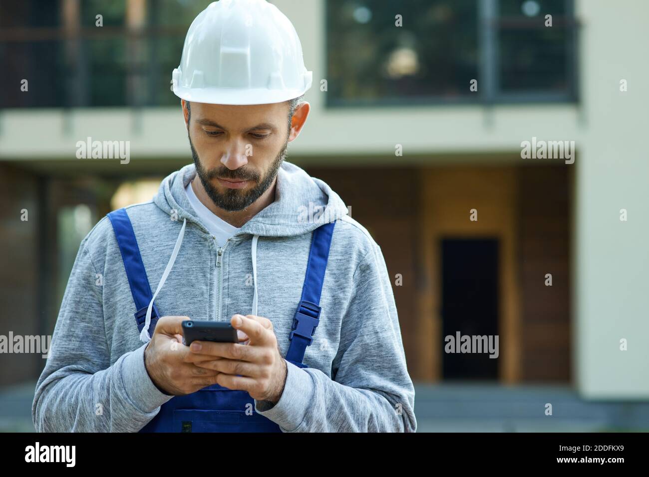 Young male builder in hardhat using smartphone, standing outdoors while working at cottage construction site. Profession, construction and building concept. Front view Stock Photo