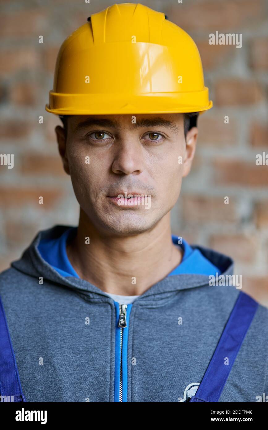 Close up portrait of handsome charismatic young male builder in hard hat looking at camera, posing on a brick wall background, working at construction site. Building, people concept Stock Photo