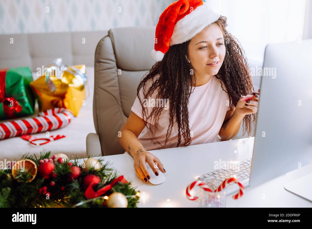 Young afro haired woman wearing santa hat shop online sitting at home in Christmas decorated room paying with bank card Stock Photo