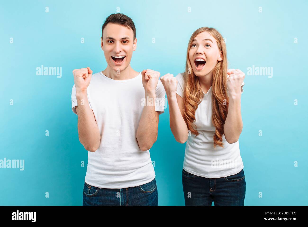 Portrait of an excited couple, a man and a woman in white t-shirts celebrating victory with fists raised, screaming, on a light blue background Stock Photo