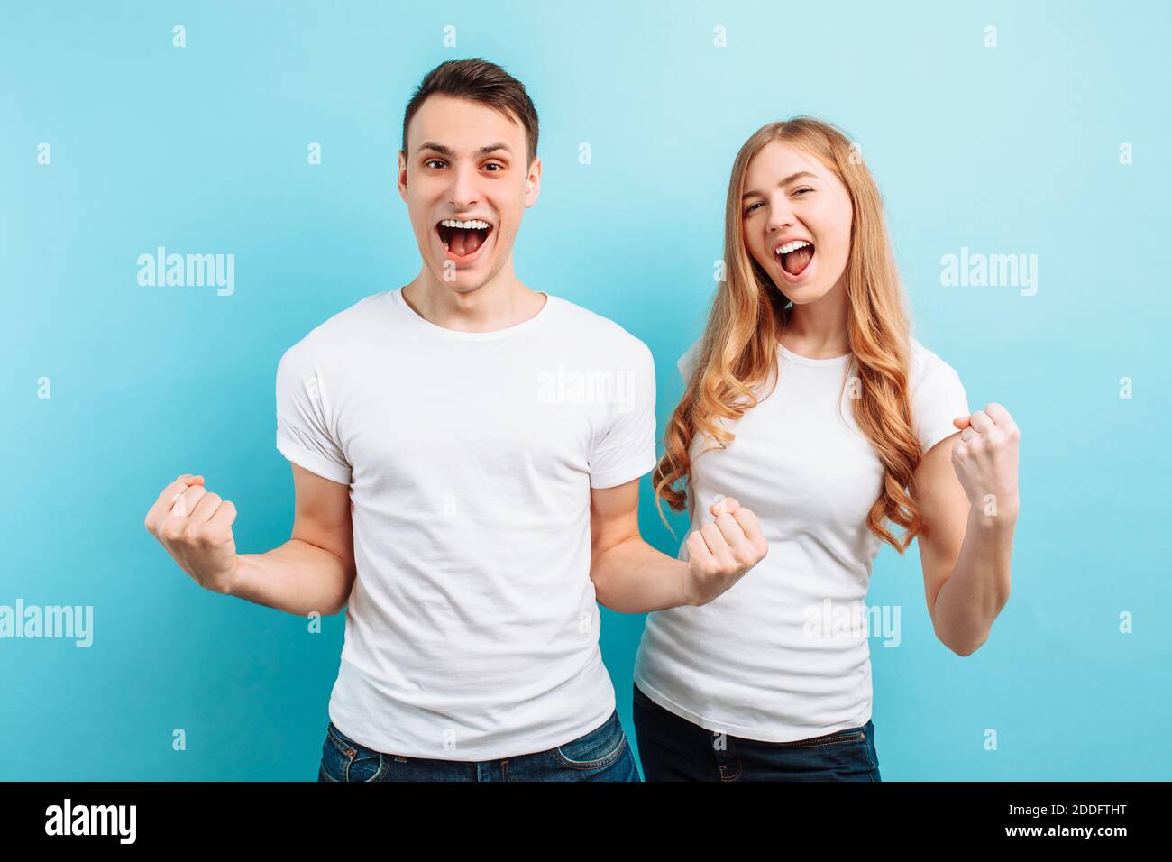 Portrait of an excited couple, a man and a woman in white t-shirts celebrating victory with fists raised, screaming, on a light blue background Stock Photo