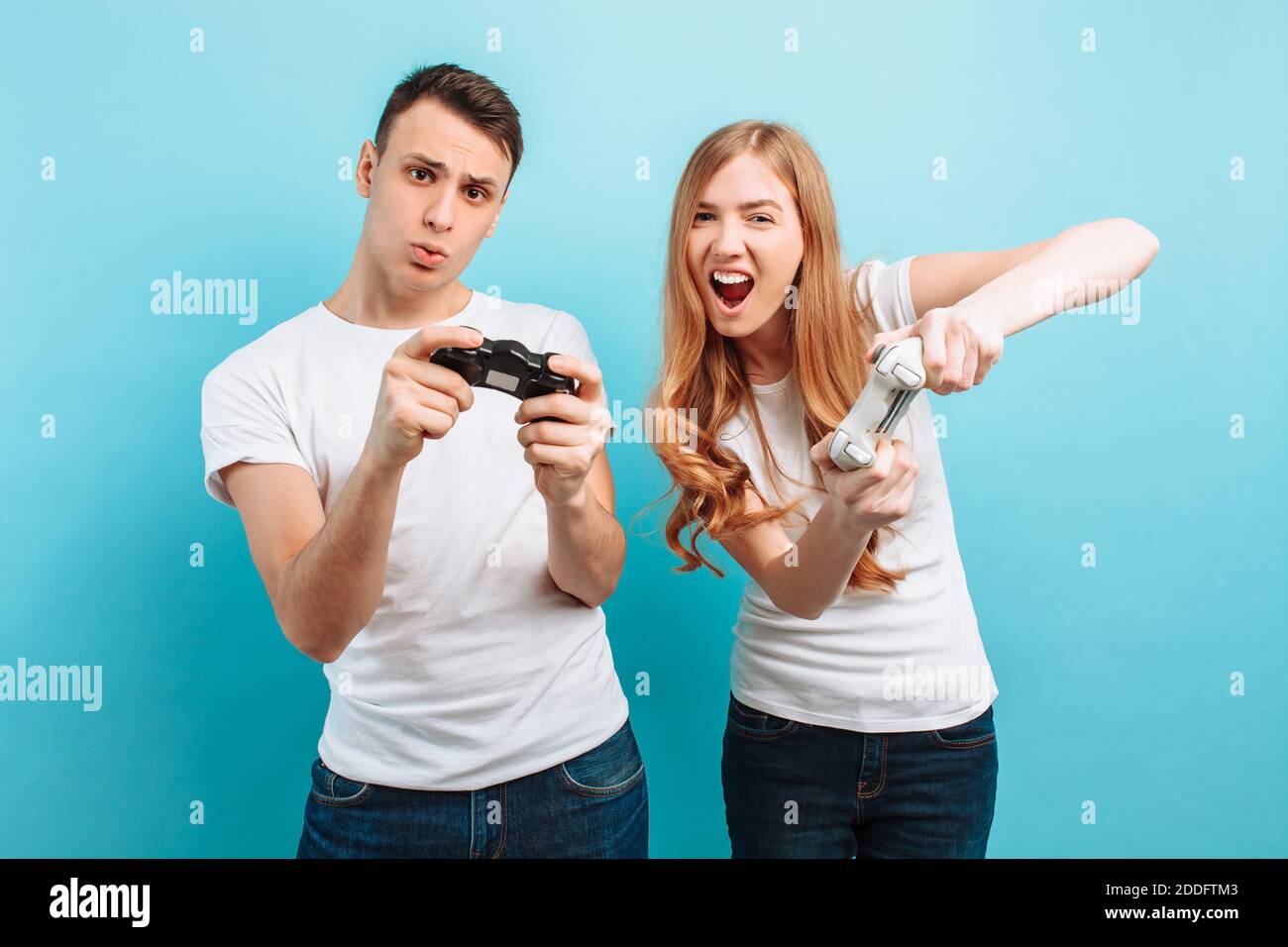 Excited young couple, a guy and a girl in white T-shirts with joysticks in their hands playing video games on a blue background Stock Photo