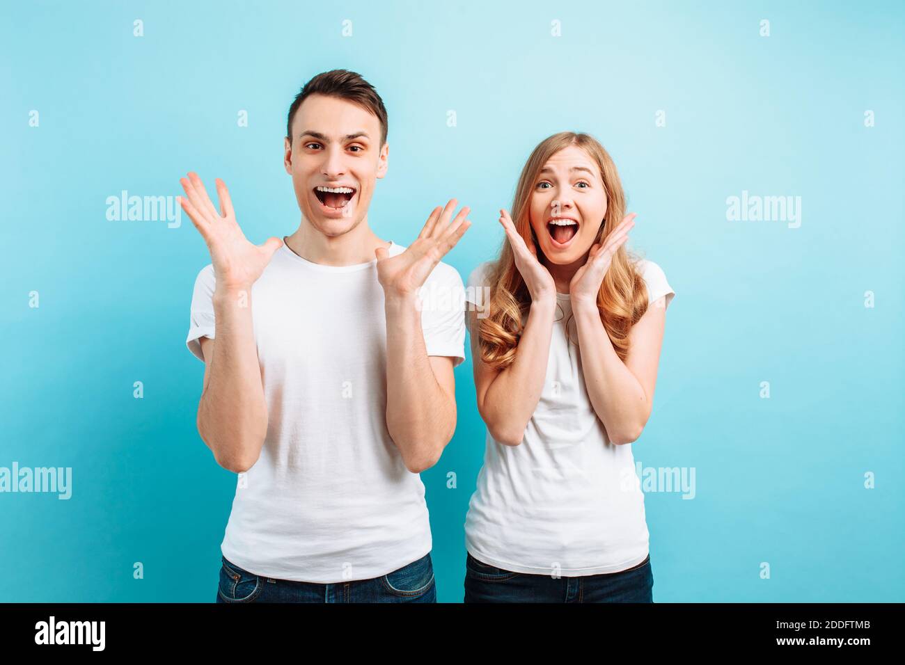 Photo of an emotional pair of a man and a woman in white T-shirts screaming with surprise or delight and raising their hands against a blue background Stock Photo