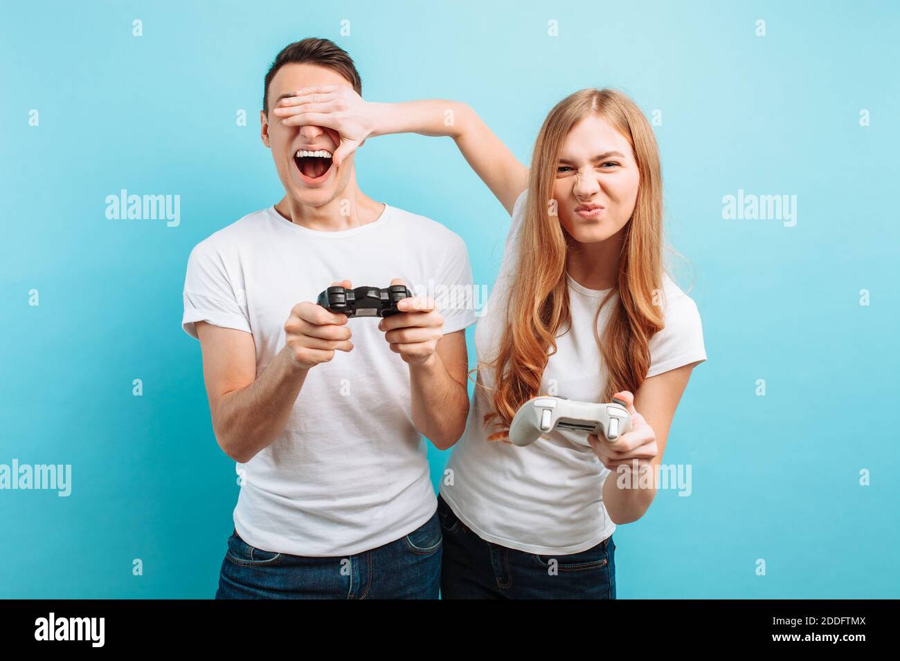 Excited young couple, a guy and a girl in white T-shirts with joysticks in their hands playing video games on a blue background Stock Photo