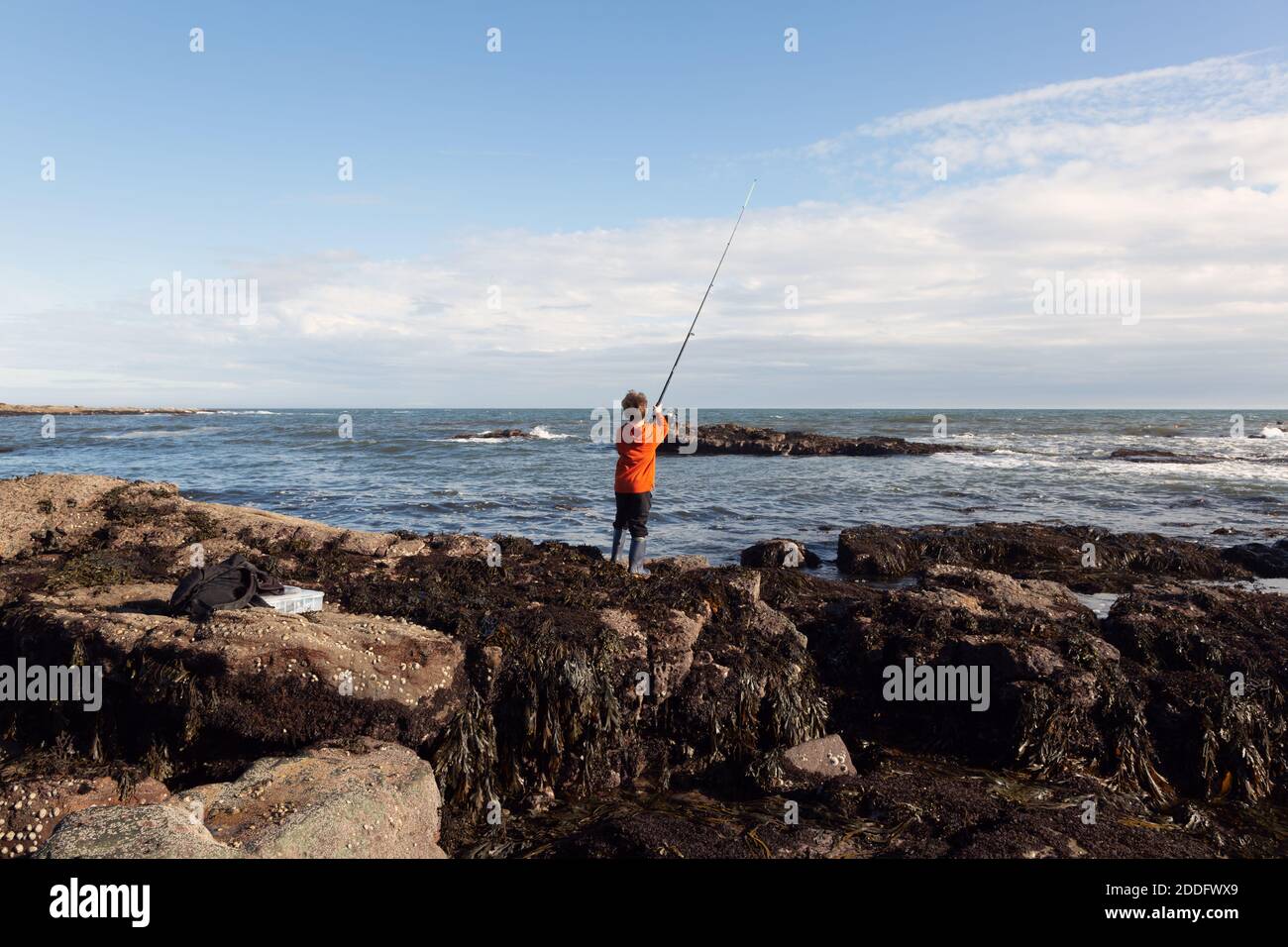 A 6yr old boy is fishing in the sea, with his tackle box and a bag on the rocks behind him. He is using a weedless lure. Stock Photo