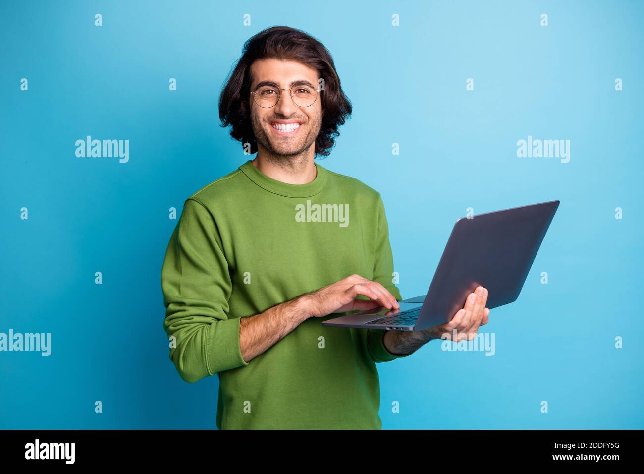 Photo of cheerful programmer guy hold netbook look camera wear glasses green pullover isolated blue color background Stock Photo