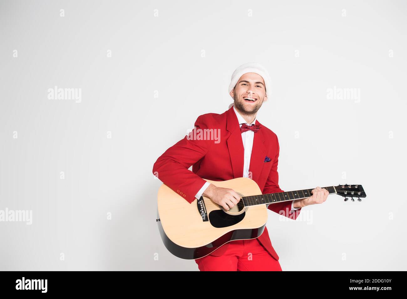 Young man in suit and santa hat smiling at camera while playing acoustic guitar on grey background Stock Photo