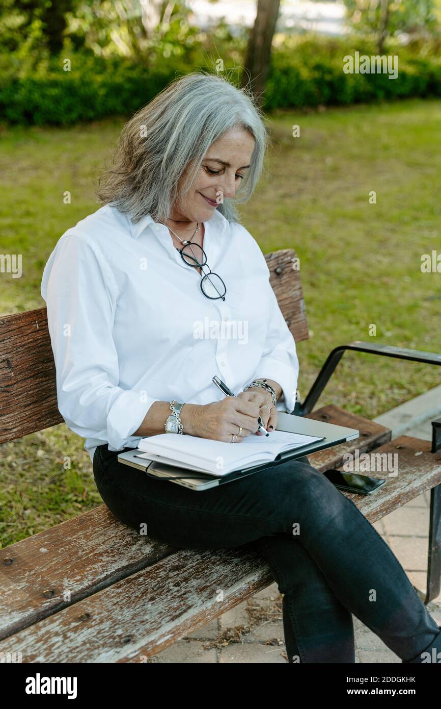 Middle aged female entrepreneur in smart casual wear sitting on bench and writing list of daily routine tasks while working remotely in park Stock Photo
