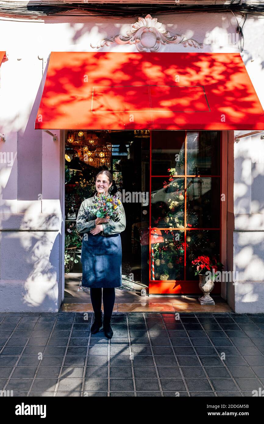 Woman holding flower bouquet standing outside shop with christmas decorative baubles and garlands in daylight looking at camera Stock Photo