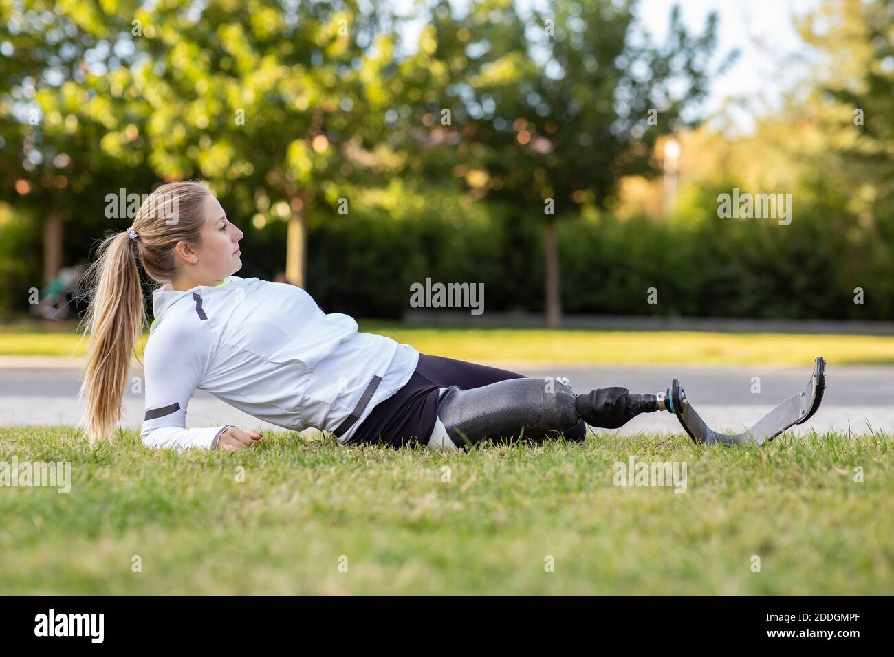 Side view of female Paralympic runner with artificial limb stretching legs during training in park while warming up and looking away Stock Photo