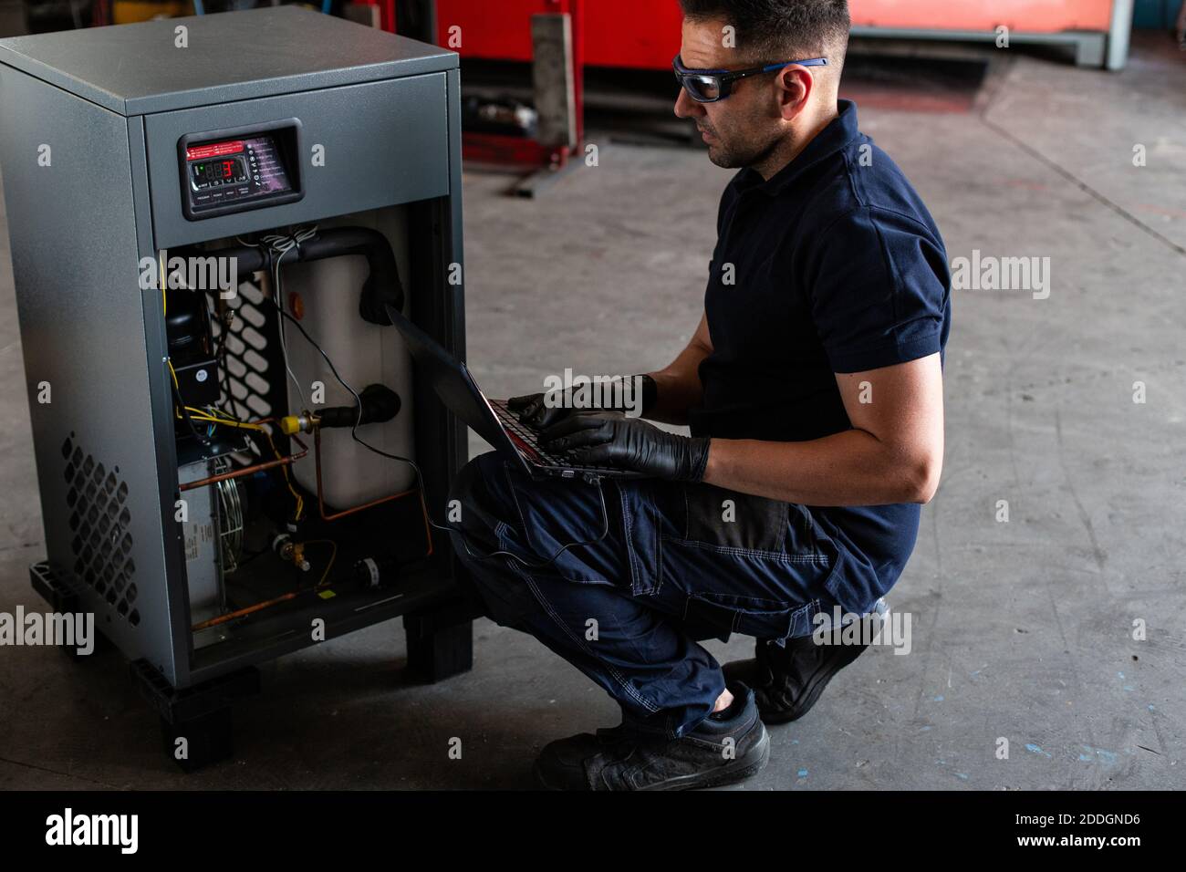 Adult man in goggles using laptop to program broken machine during work in modern workshop Stock Photo