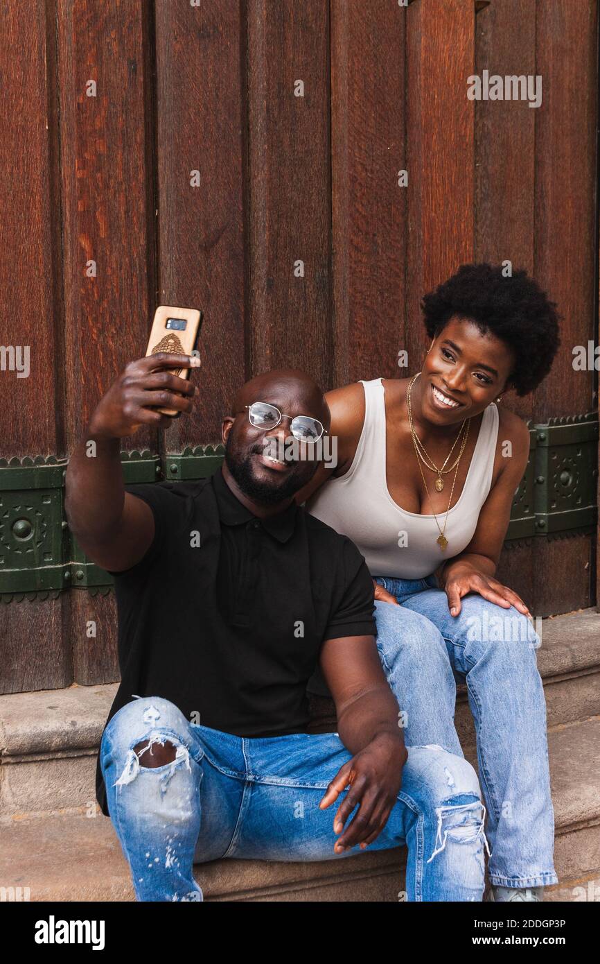 Cheerful African American couple in casual clothes sitting on stairs and taking selfie on smartphone Stock Photo