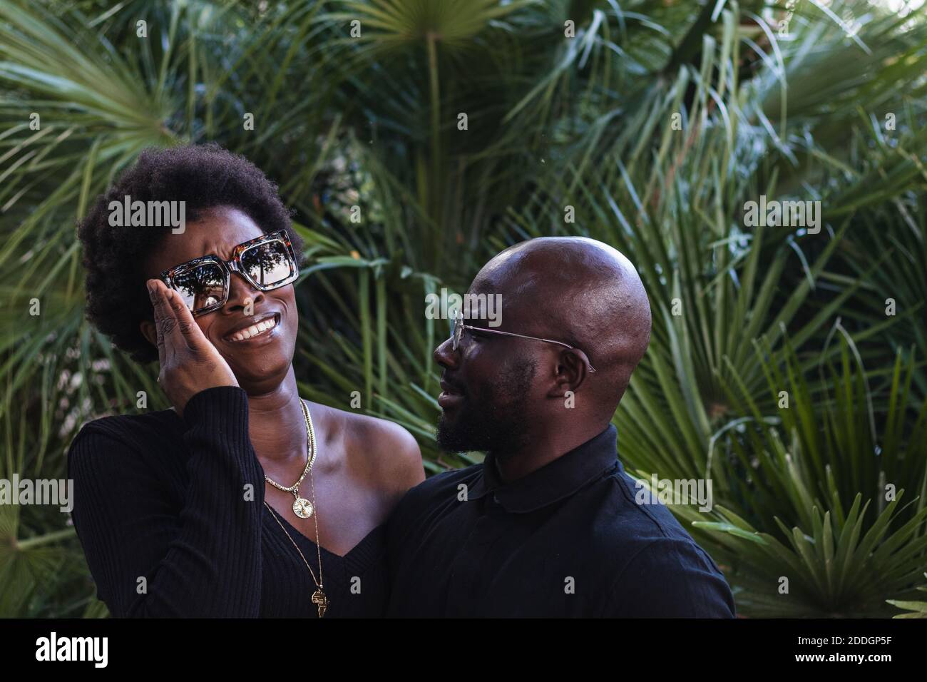 Puzzled African American female in sunglasses embracing boyfriend while resting together among green palms Stock Photo