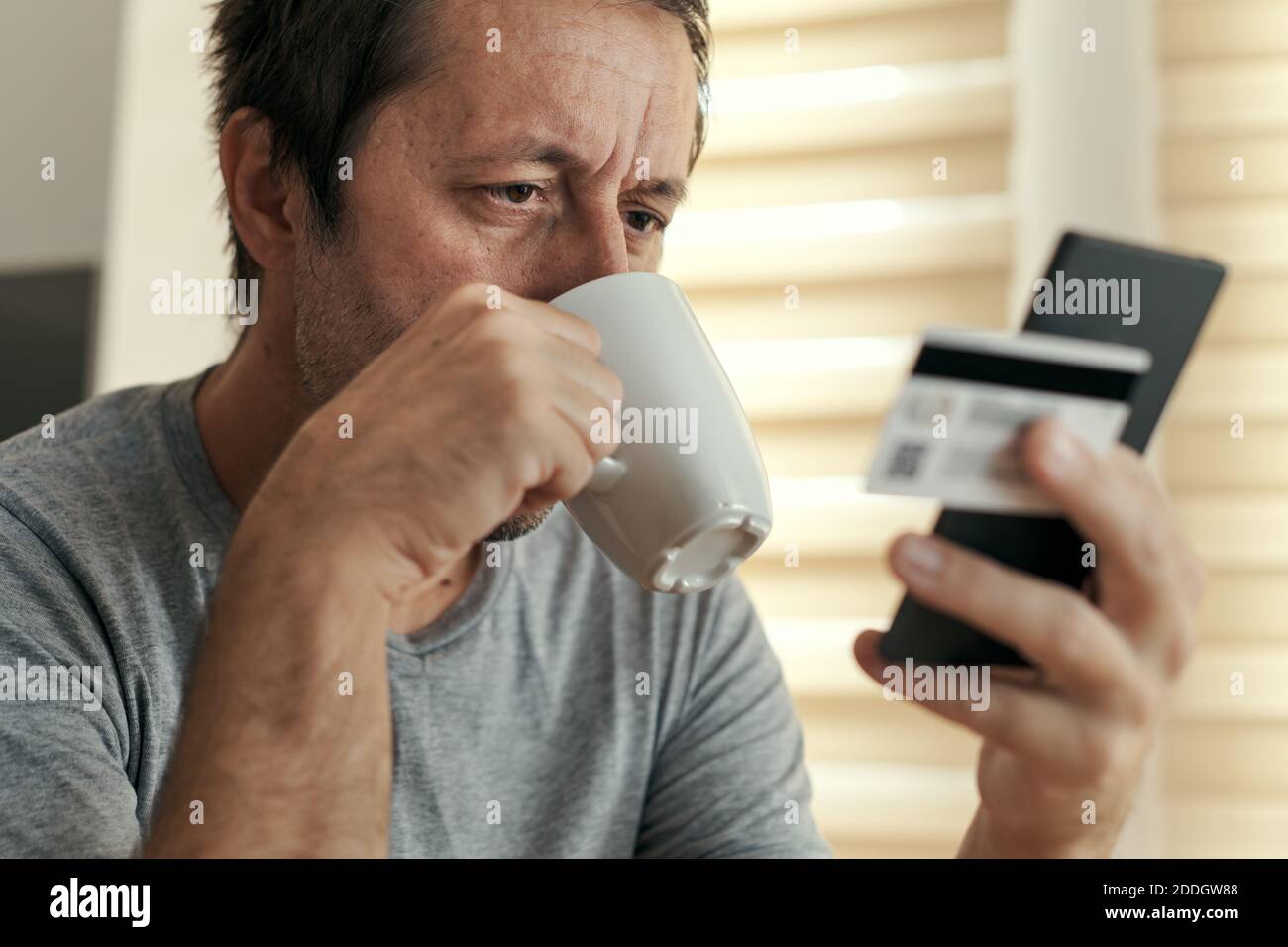 Man shopping online from home with mobile phone and credit card, adult male drinking coffee and buying on internet from his living room Stock Photo