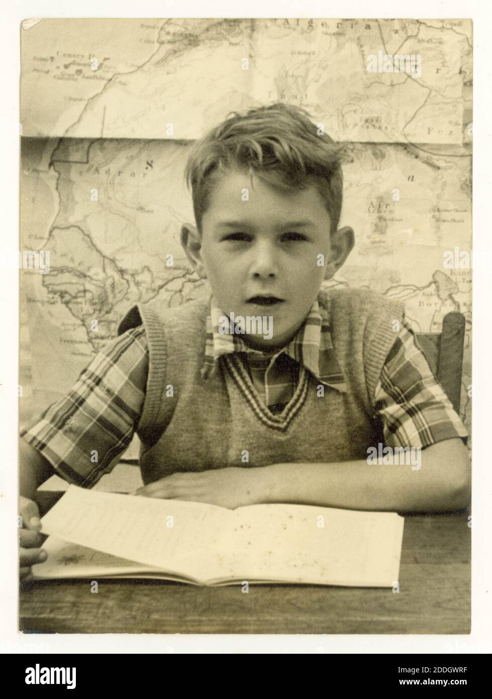 Original 1950's school photograph of school boy wearing a check shirt and tank-top sitting at a desk reading a book, map on wall behind him, U.K. Stock Photo